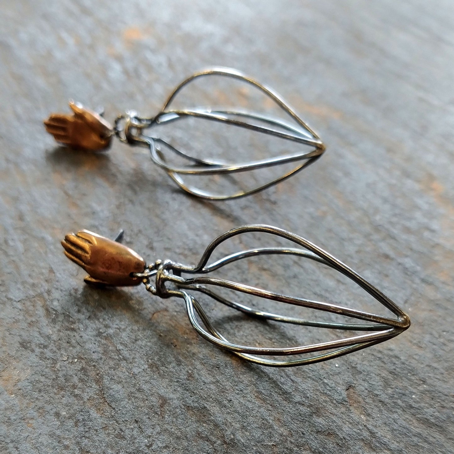 A pair of earrings featuring bronze hands above silver wire vessels with a black patina by Alice Scott