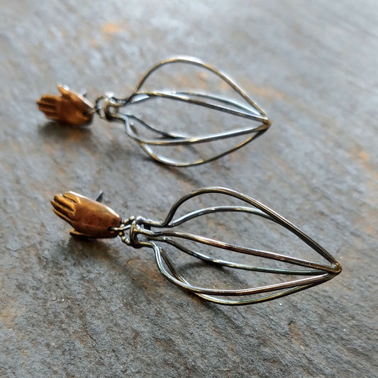 A pair of earrings featuring bronze hands above silver wire vessels with a black patina by Alice Scott