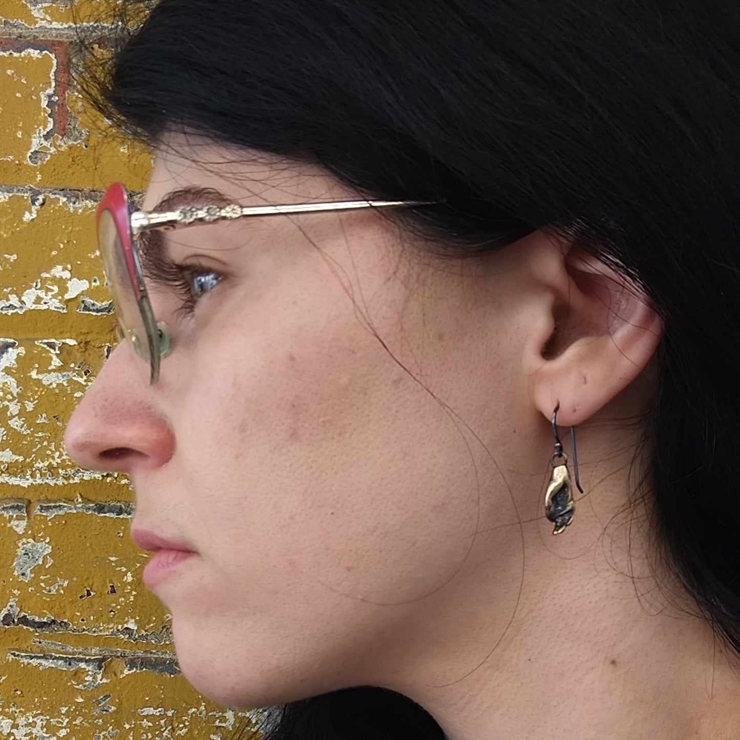 A pair of bronze earrings in the shape of hands performing a mudra, attached to oxidized silver wire hooks, set against a model ear by Alice Scott
