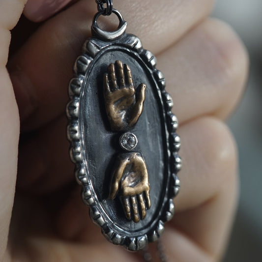 Close-up of a double hand cameo necklace with bronze hands and a sterling silver cameos background, featuring a topaz gemstone between the hands, by Alice Scott, with a hand in the backgroung