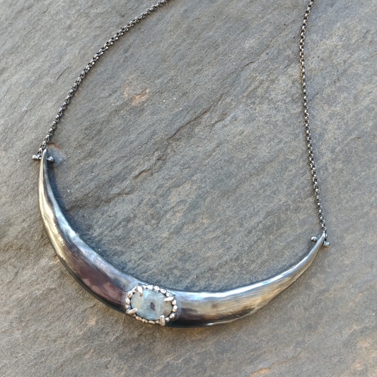 A silver crescent moon necklace with a black patina finish and a central aquamarine pendant, displayed against a textured background, by Alice Scott
