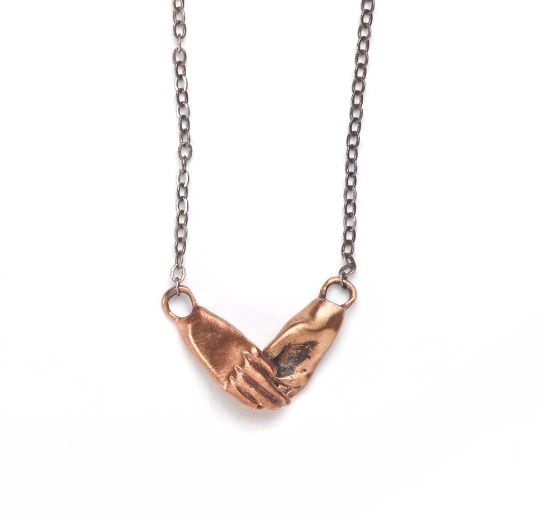 A bronze necklace featuring two hands holding each other, attached to a sterling silver chain with a sterling clasp, by Alice Scott on a white background