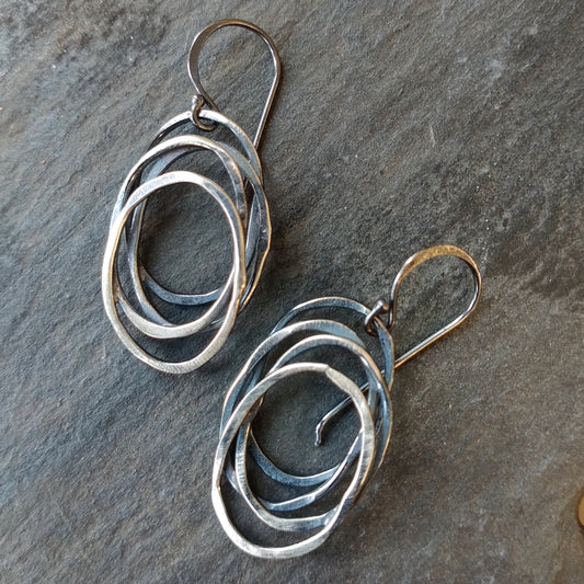 A pair of silver dangle earrings with a galaxy-inspired design, featuring multiple interwoven circles by Alice Scott on a slate background
