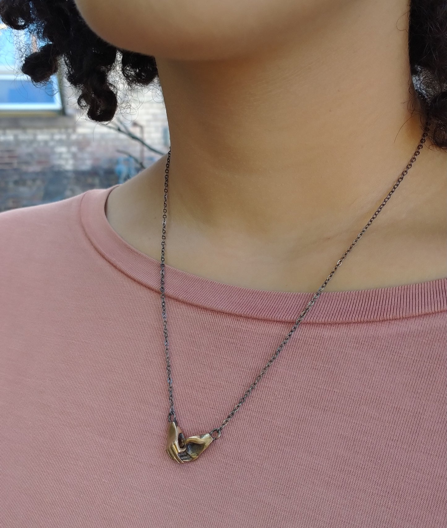 A bronze necklace featuring two hands holding each other, attached to a sterling silver chain with a sterling clasp, by Alice Scott on a model neck