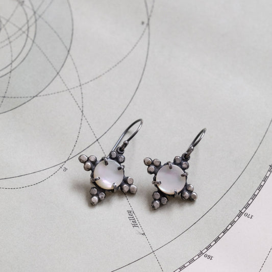 A pair of silver earrings with mother of pearl details, displayed on a piece of paper with geometric shapes, by Alice Scott.