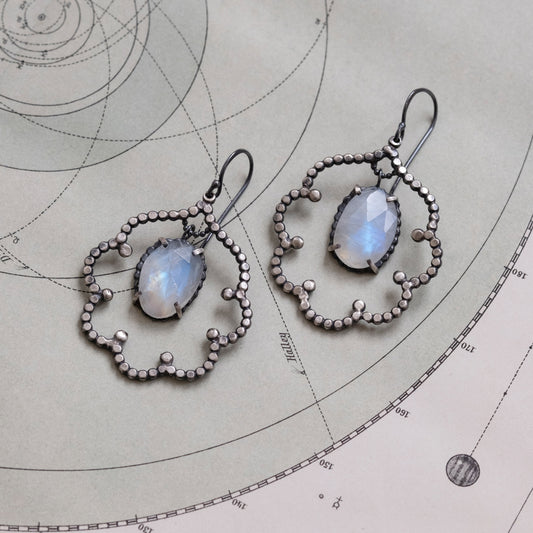 A pair of moonstone cloud earrings laid out on a flat surface with a map background. The earrings are composed of sterling silver with a black patina, featuring moonstone gemstones set within cloud-shaped outlines with intricate detailing by Alice Scott