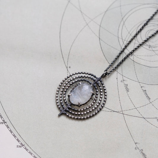A necklace with a central moonstone surrounded by a circular silver frame with intricate details, placed on a paper with geometric drawings, by Alice Scott