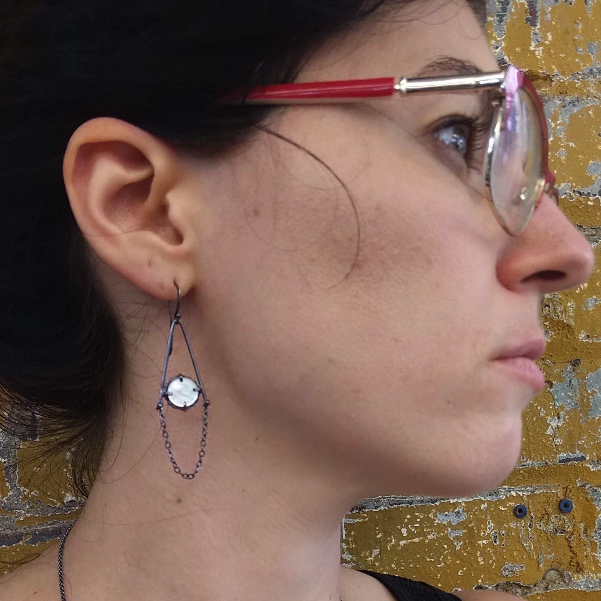 A pair of dangle earrings featuring mother of pearl circles connected to sterling silver chains, with a black patina finish, displayed against a model ear, by Alice Scott