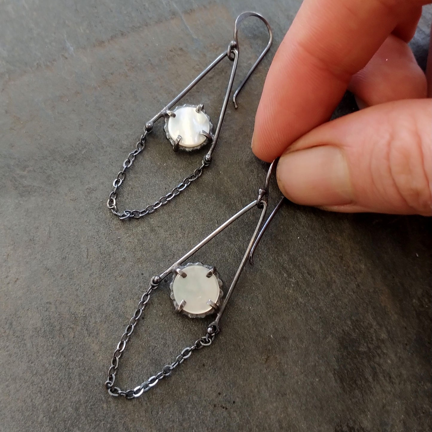 A pair of dangle earrings featuring mother of pearl circles connected to sterling silver chains, with a black patina finish, displayed against a textured grey background with a hand, by Alice Scott