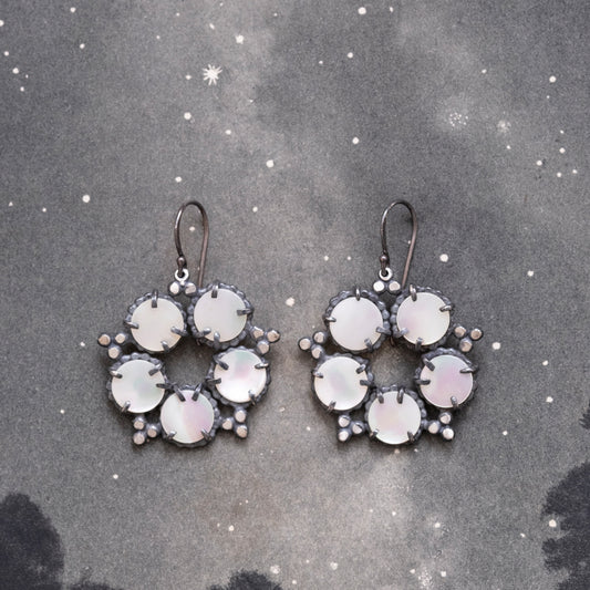 A pair of silver, mother of pearl earrings with a star and constellation design, displayed against a dark background speckled with stars by Alice Scott
