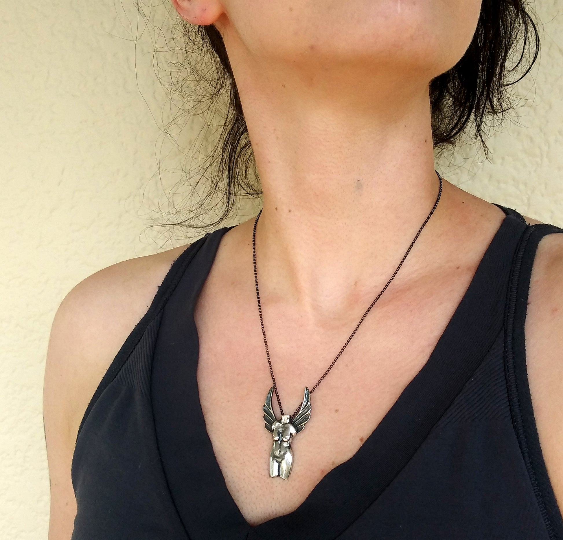A silver necklace with a detailed pendant representing the Winged Venus figure, suspended on a black patina chain on a model neck, by Alice Scott