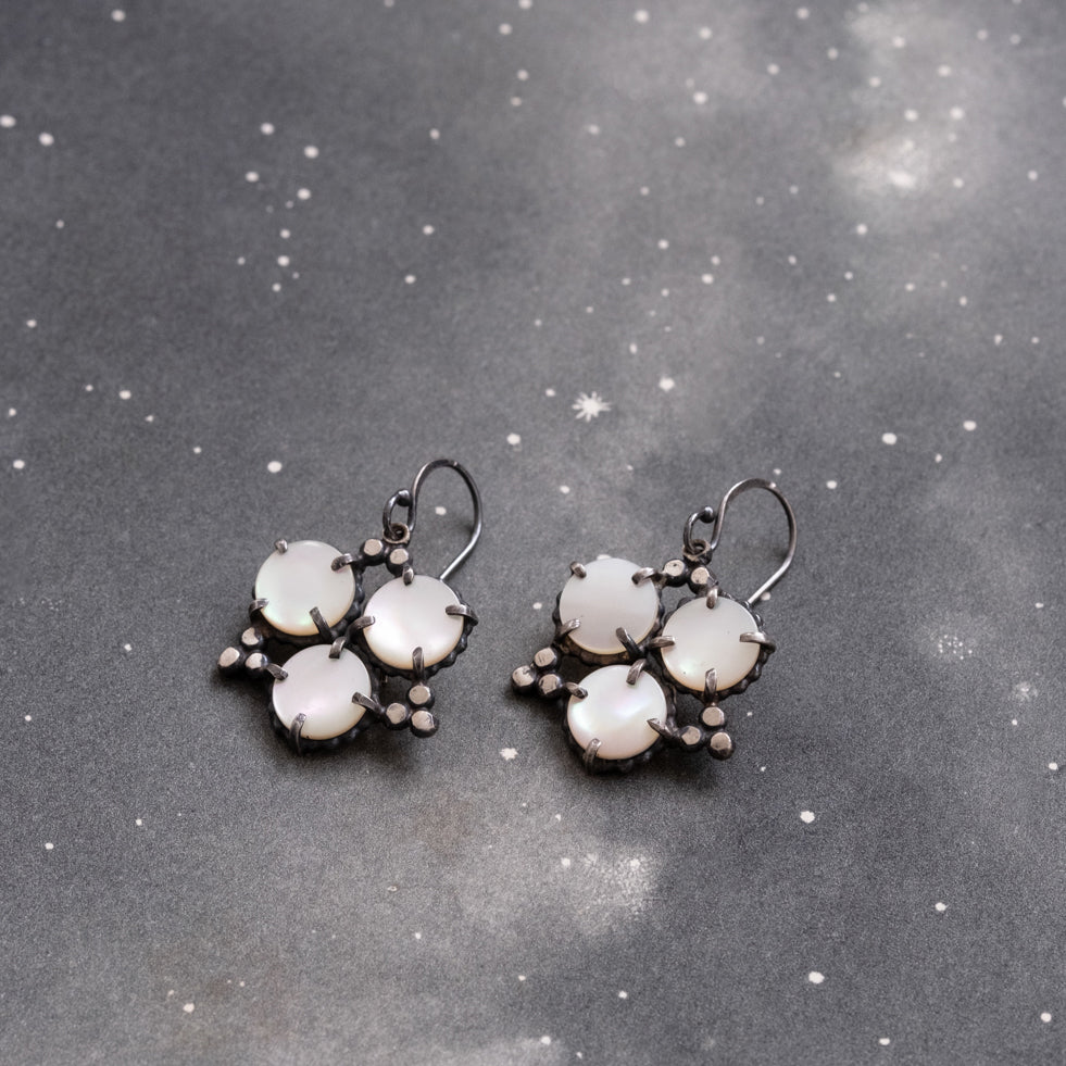 A pair of geometric pearl star earrings laid out on a textured grey surface, showcasing circular mother of pearl stones with sterling silver accents by Alice Scott