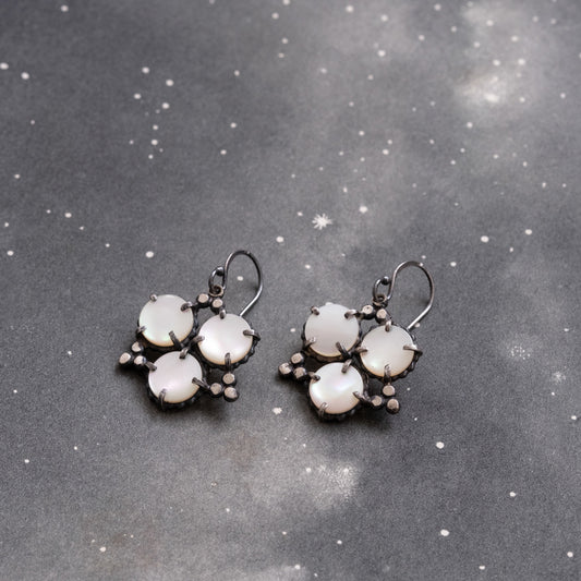 A pair of geometric pearl star earrings laid out on a textured grey surface, showcasing circular mother of pearl stones with sterling silver accents by Alice Scott